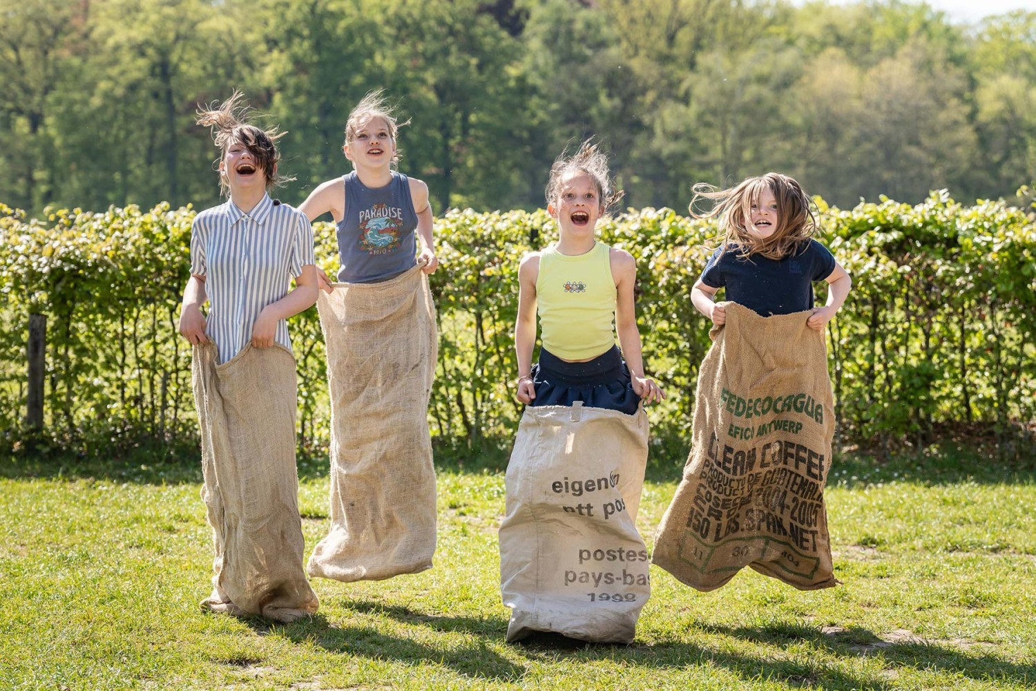 Vier kinderen springen vrolijk in jute zakken op een grasveld, omringd door groene struiken onder een heldere lucht.