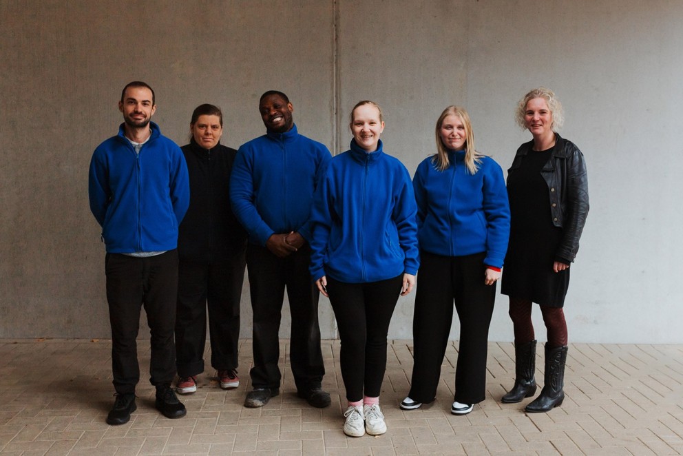 Zes mensen in blauwe sweaters staan in een rij voor een grijze wand, met een diverse samenstelling van geslachten en stijlen.