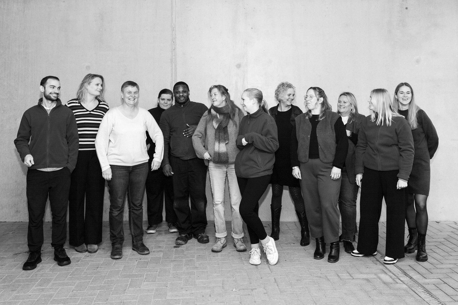 Groepsfoto van een diverse groep mensen die in een informele setting poseert, met een betonachtige achtergrond.