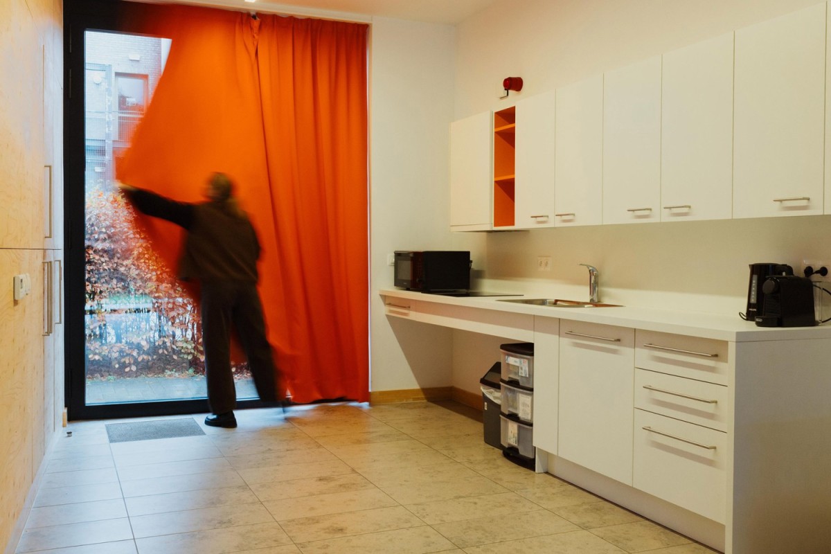 Een persoon trekt een oranje gordijn open in een moderne keukenruimte met witte meubels, een aanrecht en enkele apparaten.