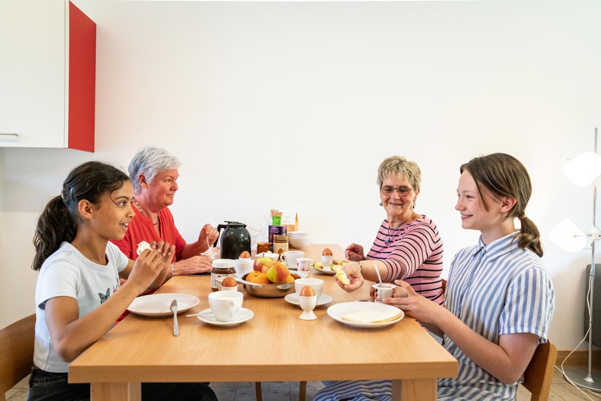 Een groep van vier mensen, waaronder twee kinderen en twee oudere vrouwen, zittend aan een eettafel met ontbijtgerechten zoals eieren, fruit en brood.