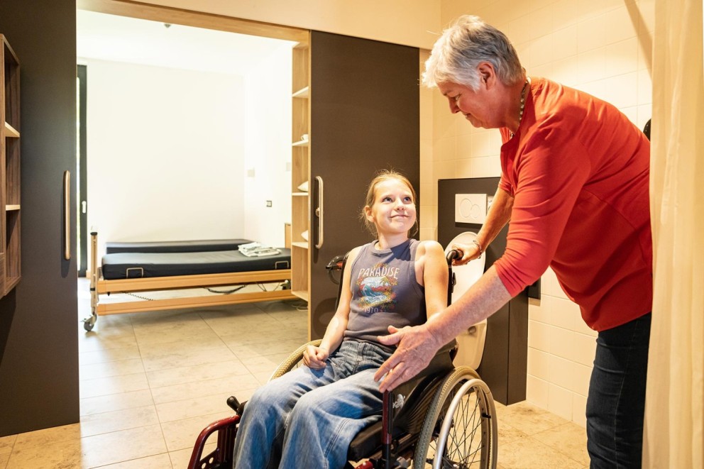 Een jonge meid in een rolstoel lacht terwijl een volwassene haar begeleidt in een moderne, lichte ruimte met een bed op de achtergrond.
