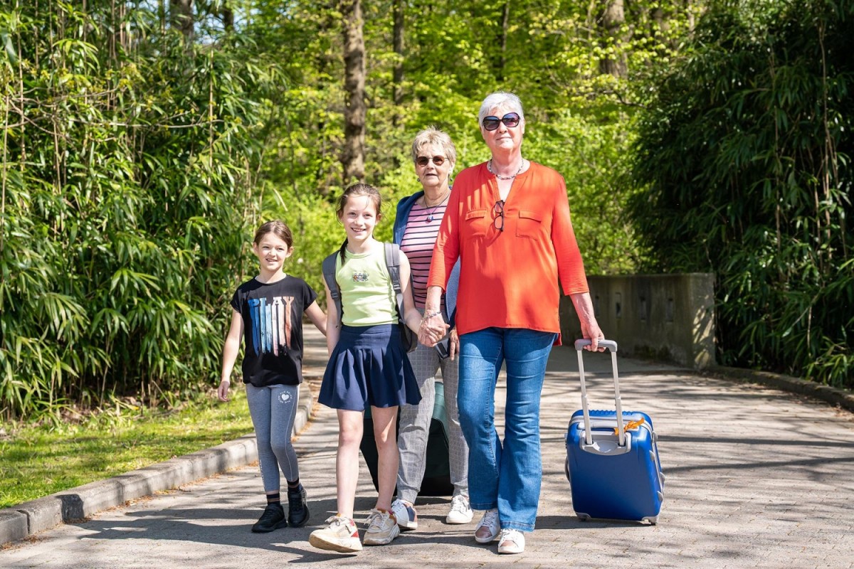 Een jonge familie, bestaande uit twee meisjes en twee vrouwen, wandelt vrolijk met bagage langs een groene, bomenrijke omgeving.