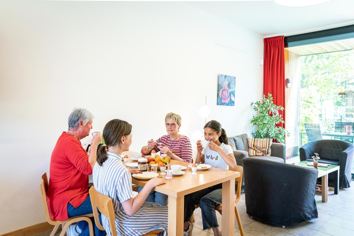 Een groep van vier mensen zit aan een tafel in een woonkamer en geniet van een ontbijt met verschillende gerechten, fruit en drankjes. In de achtergrond is een grote raampartij en een plant zichtbaar.