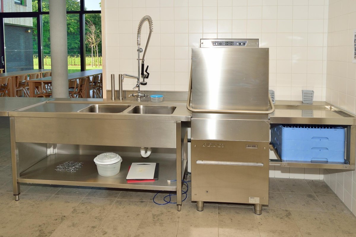 Stainless steel keuken met dubbele spoelbak, afwasmachine en werkoppervlak, gelegen in een lichte ruimte.
