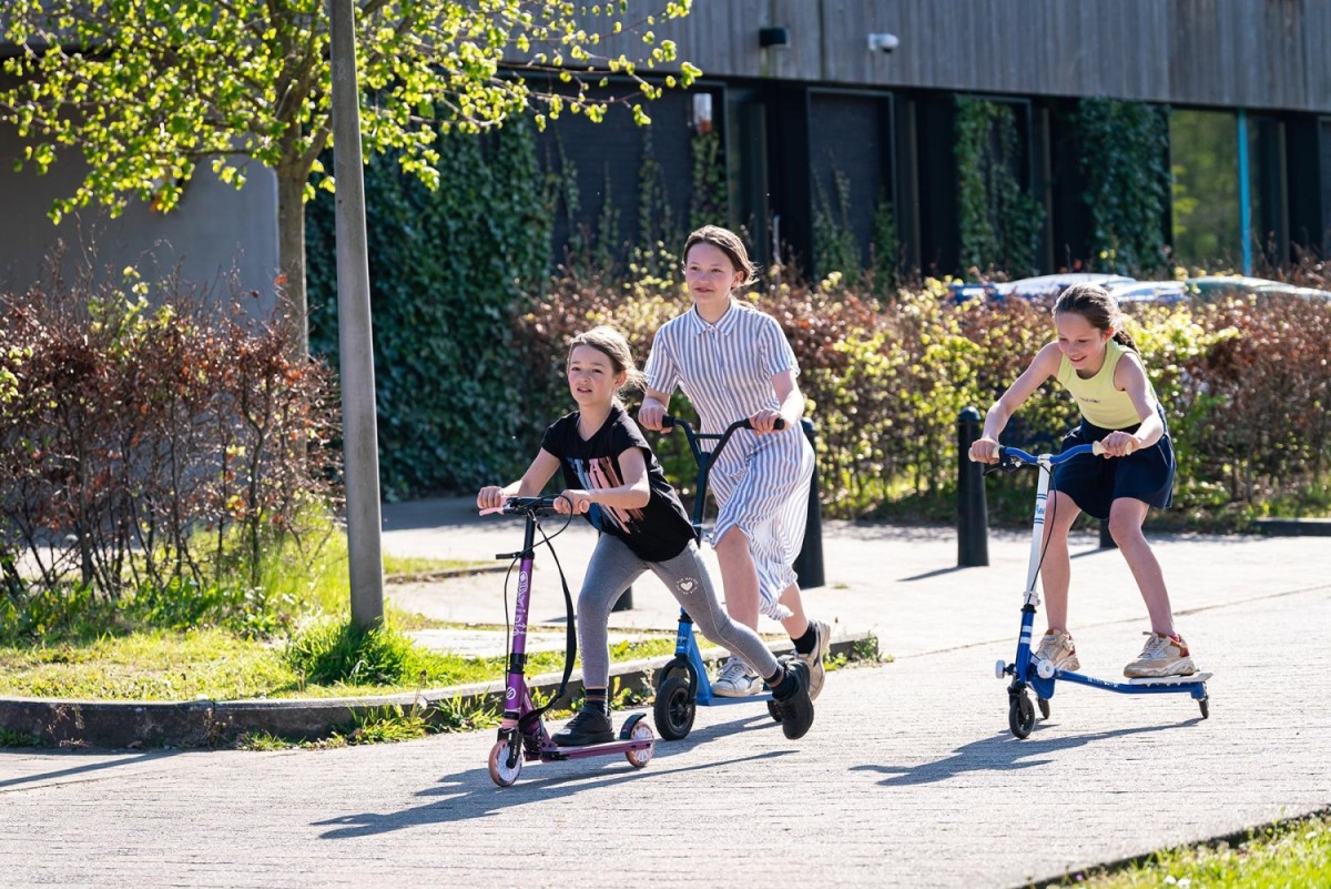 Drie kinderen rijden op stepjes in een zonovergoten omgeving, omringd door groen en planten.