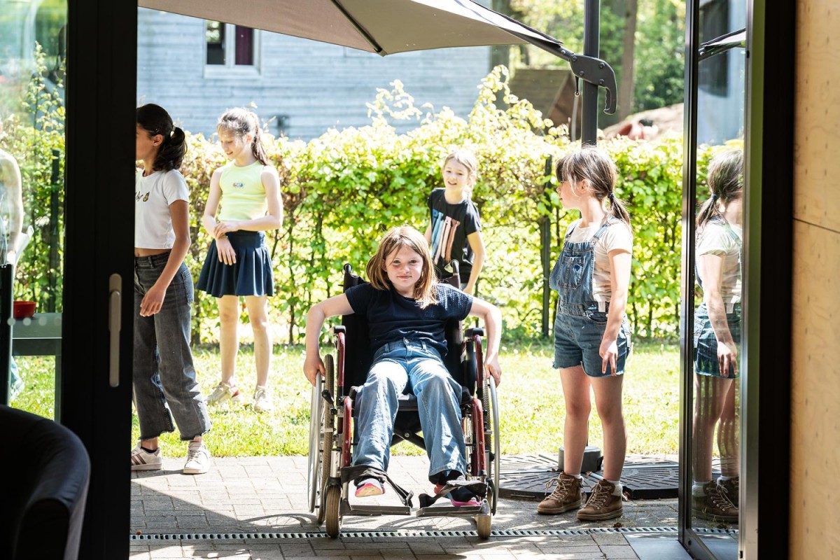 Een groep kinderen speelt buiten, terwijl een meisje in een rolstoel op de voorgrond zit en naar de camera kijkt.