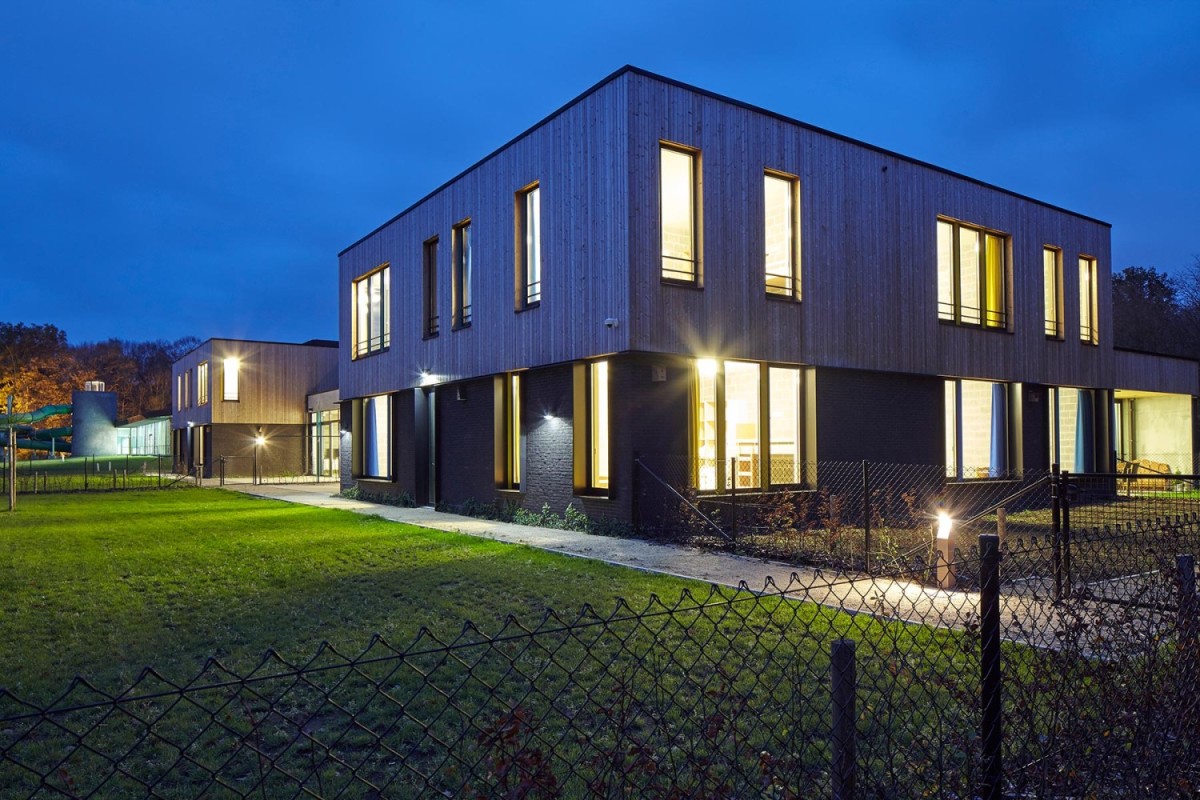 Moderne gebouwen met houten gevels, verlicht in de avond, omringd door een grasveld en een hek.