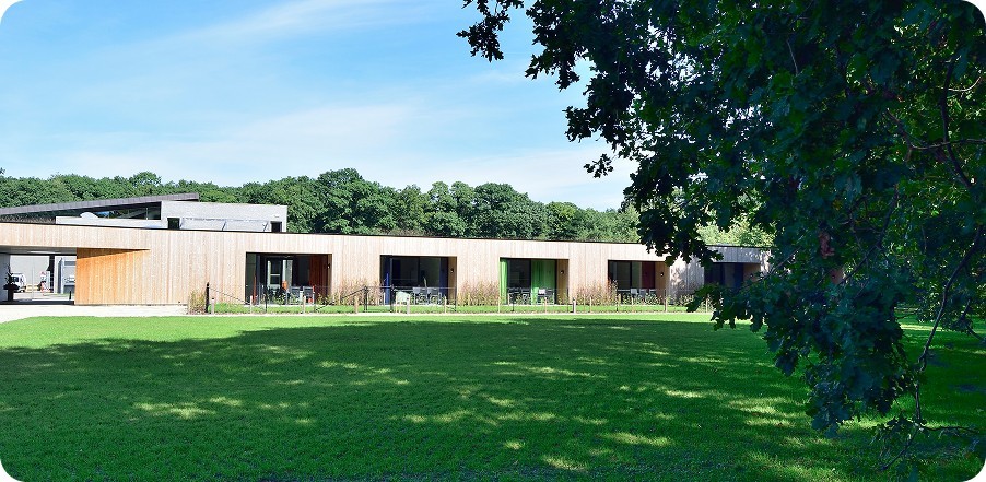 Moderne houten gebouw in het groen met grasveld en bomen op de achtergrond.