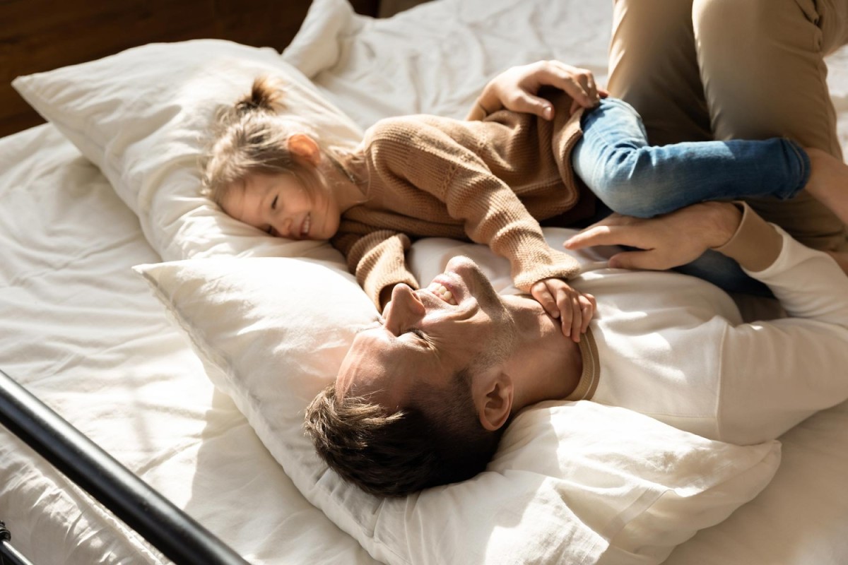 Een vader ligt op een bed met kussens terwijl hij lachend interactie heeft met zijn jonge dochter, die ook lacht en zich naast hem bevindt.