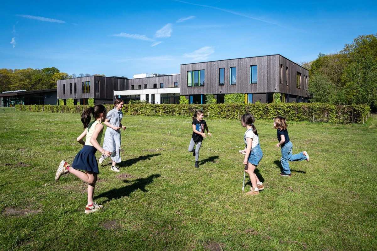 Groep kinderen renten op een groen veld voor een modern gebouw onder een blauwe lucht.
