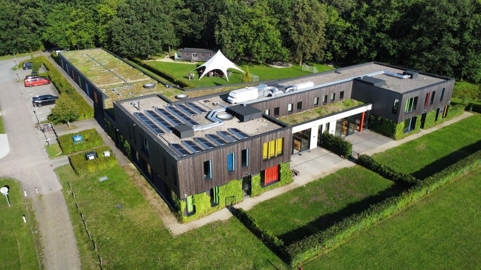 Een modern gebouw met een groen sedumdak en zonnepanelen, omringd door gras en struiken, met een witte tent in de nabijheid en parkeerplaatsen aan de zijkant.