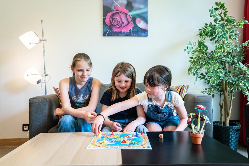 Drie kinderen zitten op een bank en spelen een bordspel, met een kleurrijk spelbord voor hen en een lamp en een plantenpot op de achtergrond.