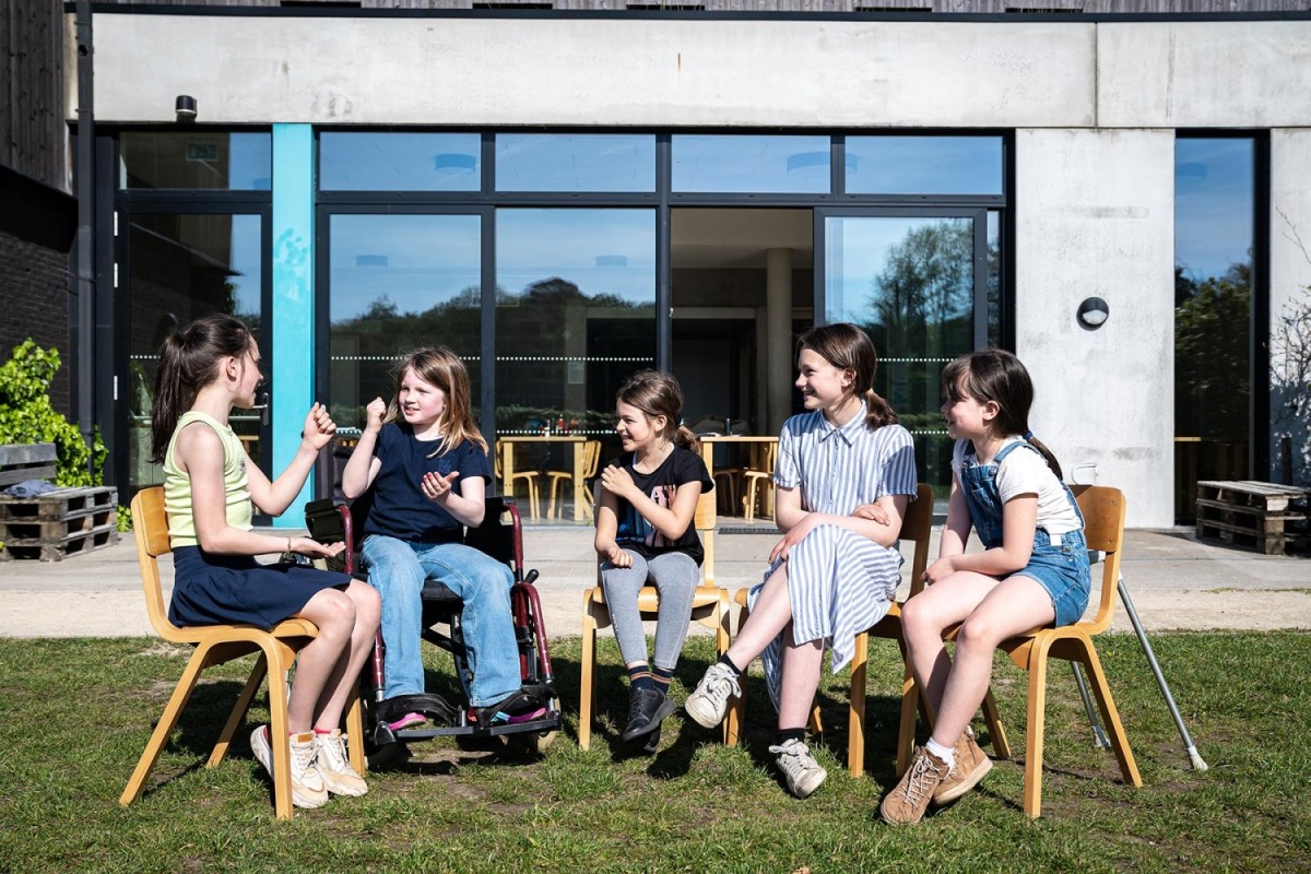 Vijf kinderen zitten op een grasveld, met &eacute;&eacute;n meisje in een rolstoel, terwijl ze met elkaar praten en lachen. Achtergrond toont een moderne gebouw met grote ramen.