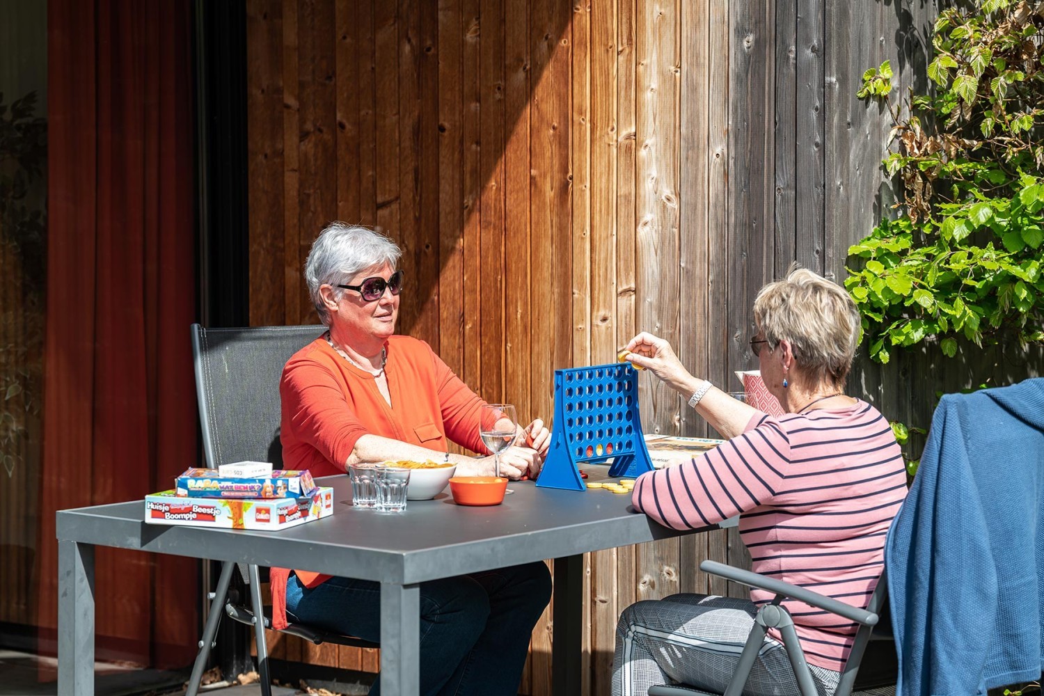 Twee vrouwen zitten aan een tafel buiten, genieten van snacks en spelen een bordspel onder de zon, met houten muren als achtergrond.