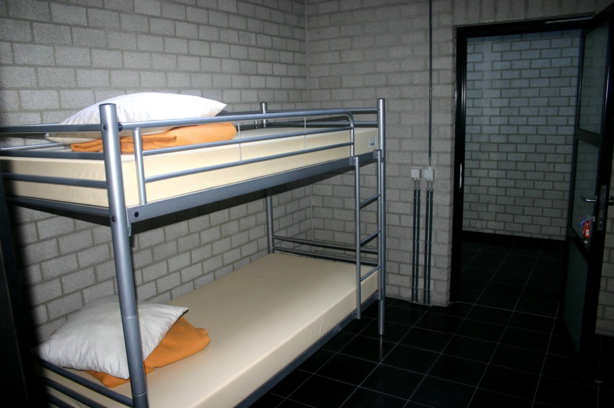 Een eenvoudige slaapzaal met een stapelbed, een wit kussen en een oranje dekentje, tegen een muur van betonblokken en met een open deur naar een andere kamer.