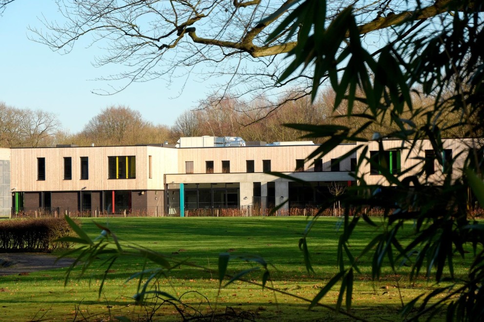 Moderne, lage gebouwen met houten gevels en grote ramen, omgeven door een groene grasvlakte en bomen op de achtergrond.