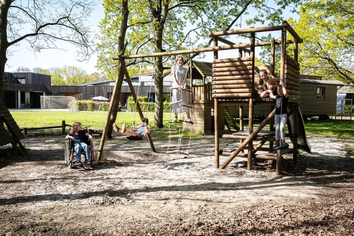 Kinderen spelen op een speelplaats met een schommel en een houten speelhuisje in een groene omgeving.
