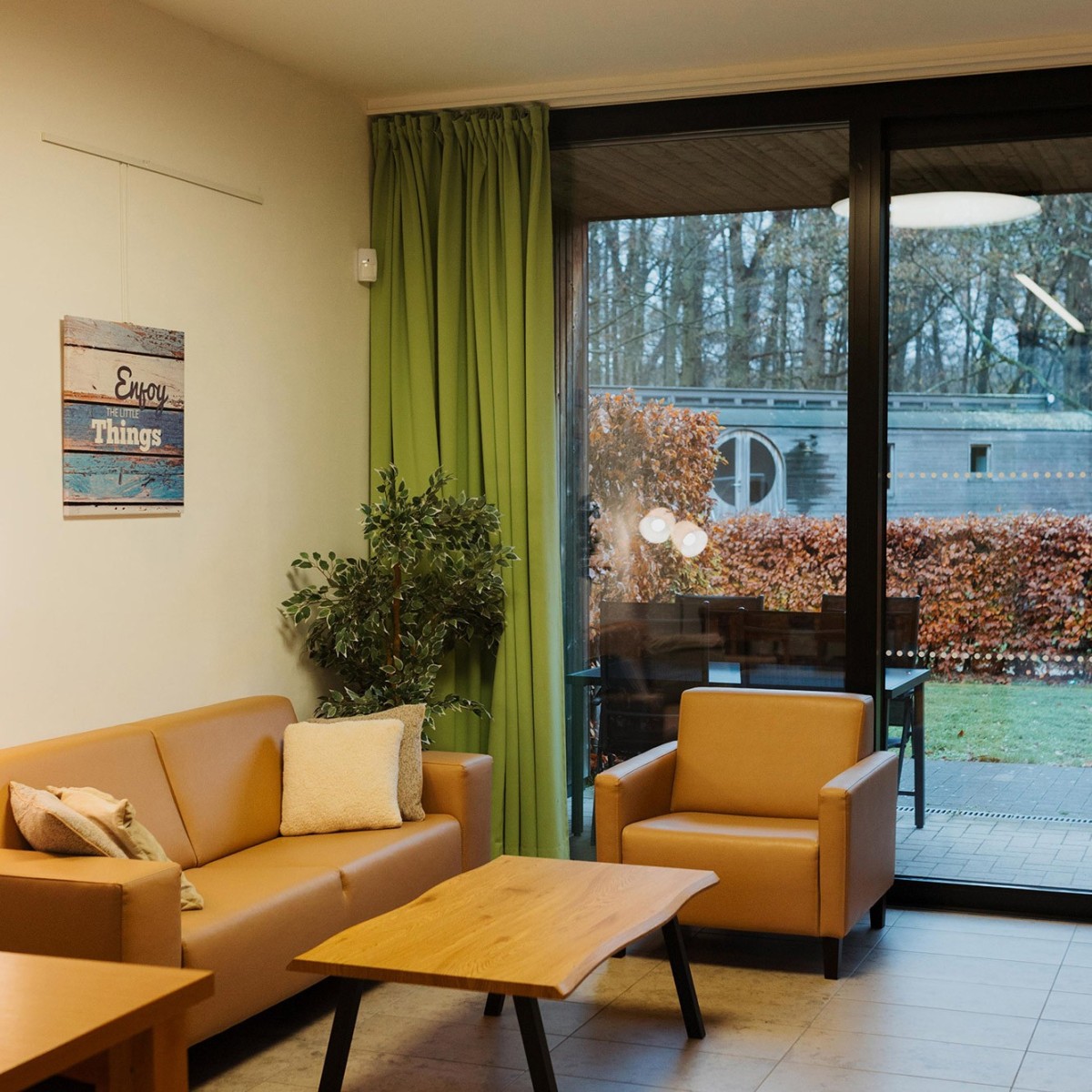 Een moderne woonkamer met een leren bank en fauteuils, een houten salontafel, groene gordijnen en een uitzicht op een tuin achter grote glazen deuren. Een afbeelding met de tekst "Enjoy the little things" hangt aan de muur.