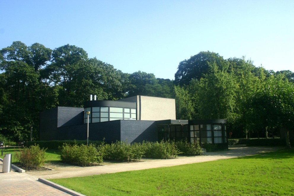 Moderne kubusvormige woning met grote ramen, omgeven door groen en bomen, onder een blauwe lucht.
