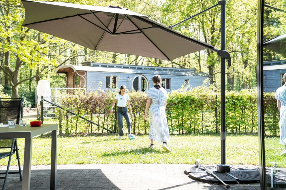Twee meisjes spelen met een voetbal in een groene tuin onder een parasol, met een houten woning op de achtergrond.