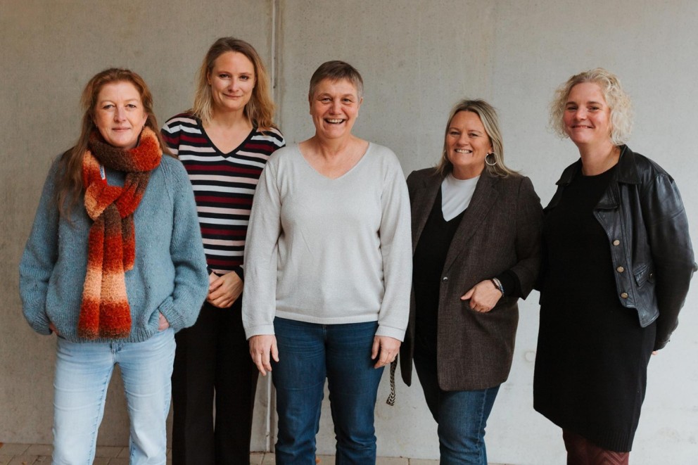 Vijf vrouwen staan naast elkaar en kijken naar de camera, met een neutrale achtergrond.