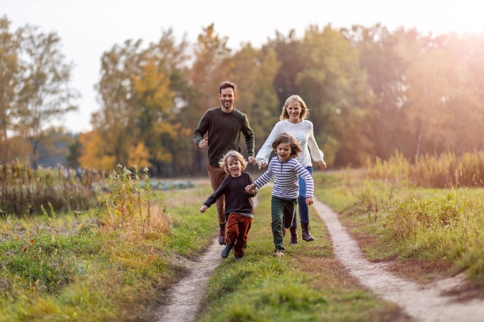 Een gezin met twee volwassenen en twee kinderen rent samen over een pad in een natuurlijke omgeving met herfstkleuren.