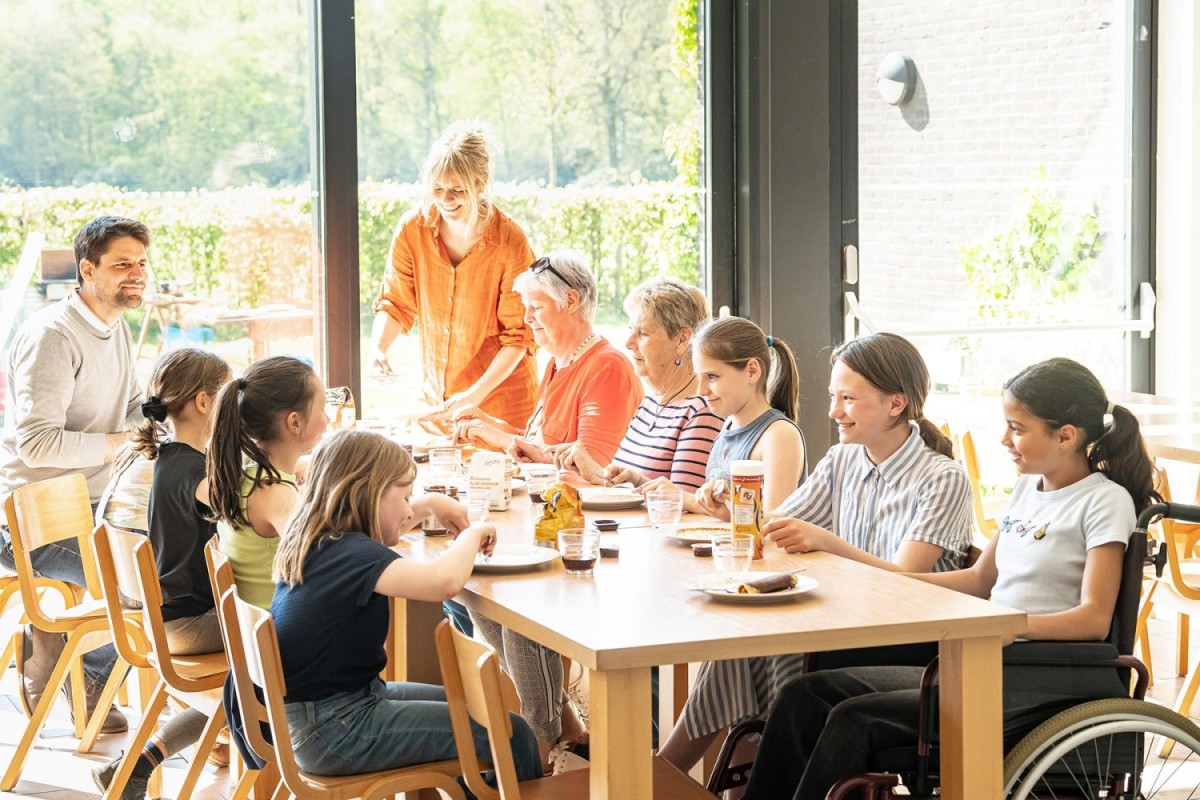 Een groep mensen, bestaande uit volwassenen en kinderen, zit aan een tafel in een lichte ruimte en geniet van een maaltijd samen. Een vrouw in een oranje blouse serveert eten, terwijl de anderen aan tafel lachen en met elkaar communiceren.