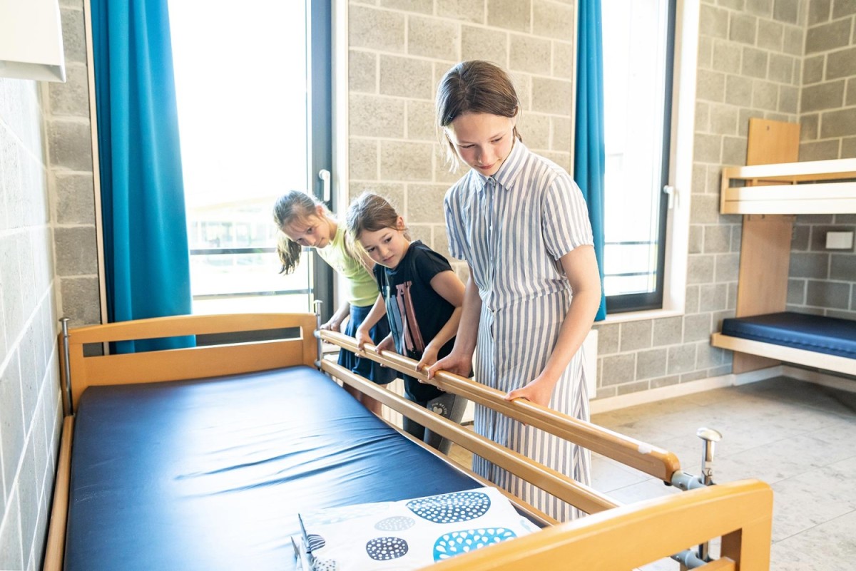Drie meisjes zijn bezig met het opmaken van een bed in een lichte kamer met grote ramen en blauwe gordijnen.