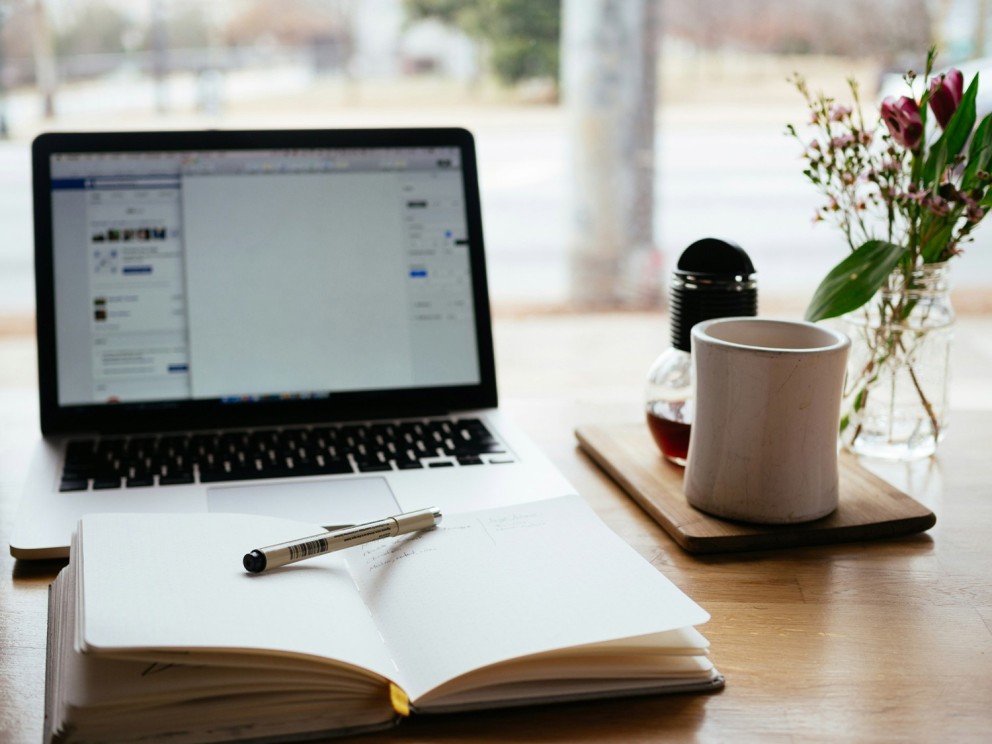 Een laptop met een blanco scherm, een notitieboek met een pen erop, een witte mok en een vaas met bloemen, alles op een houten tafel met een uitzicht op de straat.