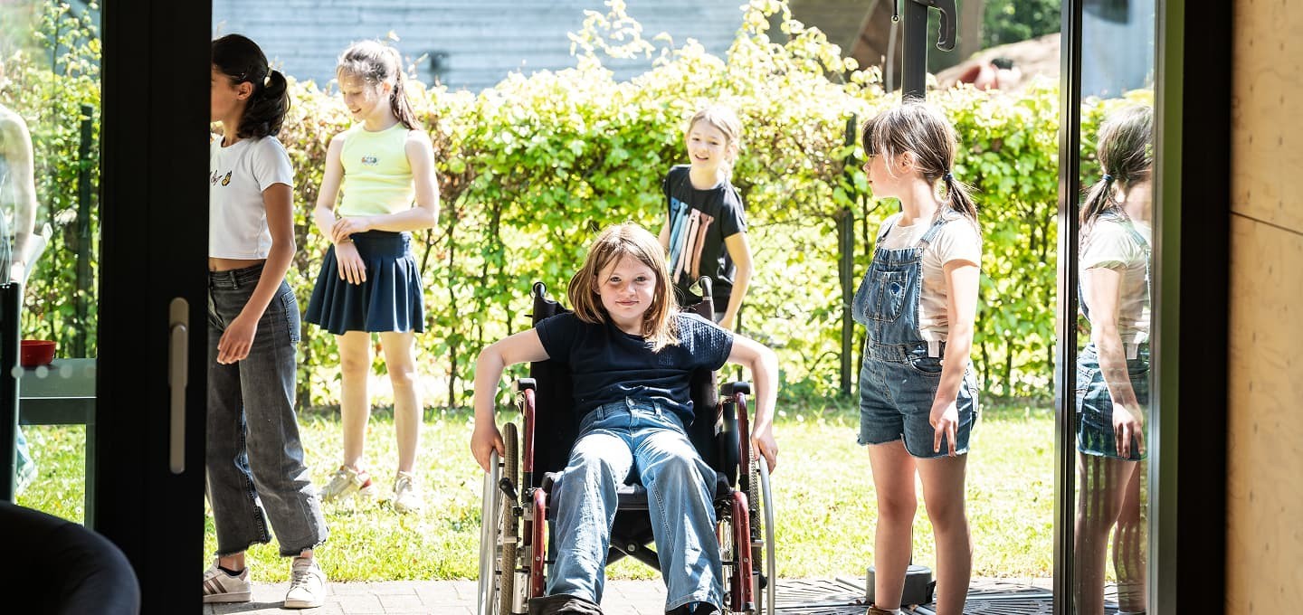 Een groep kinderen, waaronder een meisje in een rolstoel, speelt buiten op een zonnige dag, met een groene achtergrond van struiken en bomen.