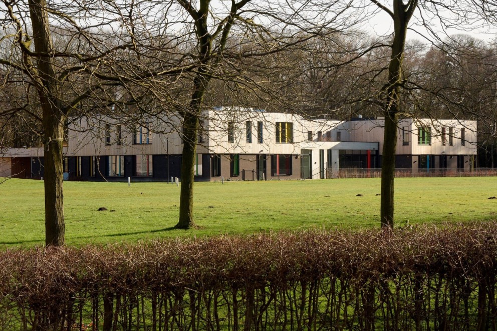 Moderne gebouwen zichtbaar achter een heg en bomen, met een grasveld ervoor.