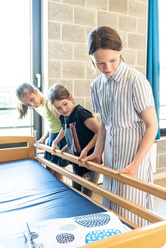 Drie kinderen in een kamer met een bed maken het bed op, terwijl ze samenwerken en naar de dekens kijken.