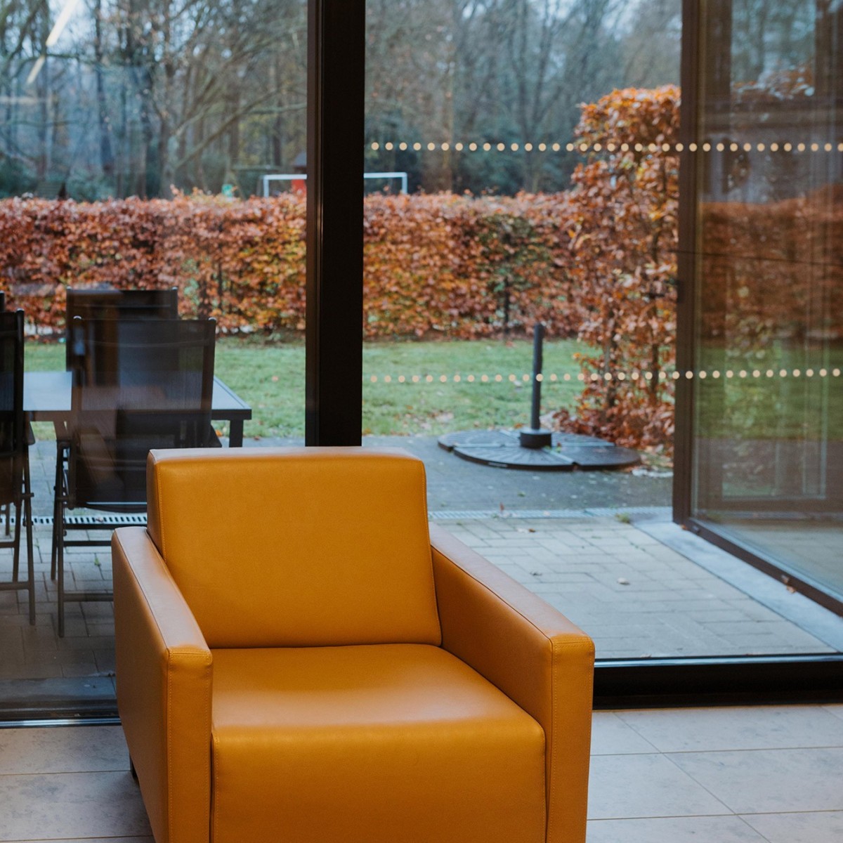 Een gele leren stoel staat naast een grote glazen wand, met uitzicht op een tuin met herfstachtige bladeren en een eettafel met stoelen op de achtergrond.