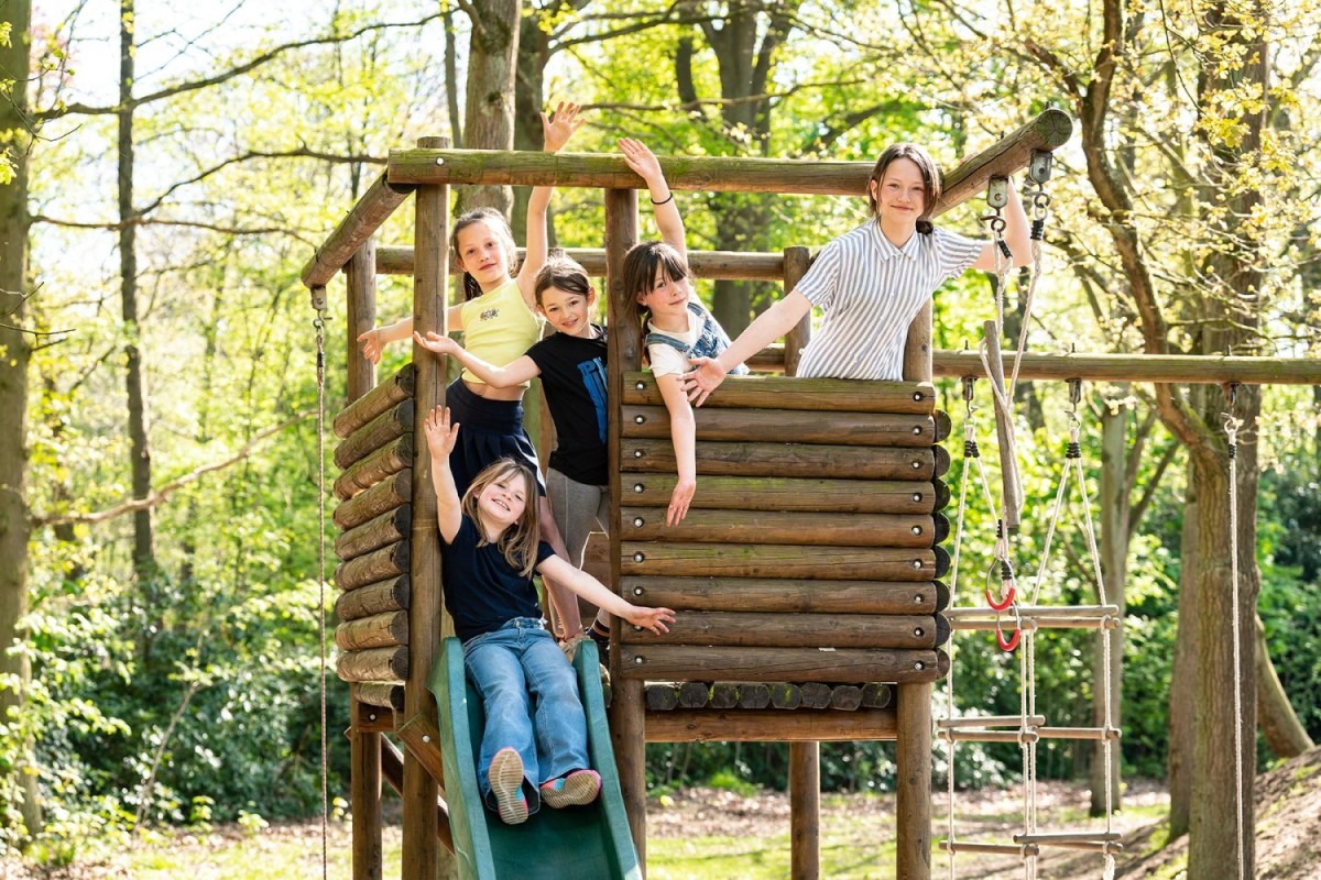 Vijf kinderen in een speeltuin, lachend en poserend op een houten speelstructuur met een glijbaan, omringd door groene bomen en zonlicht.