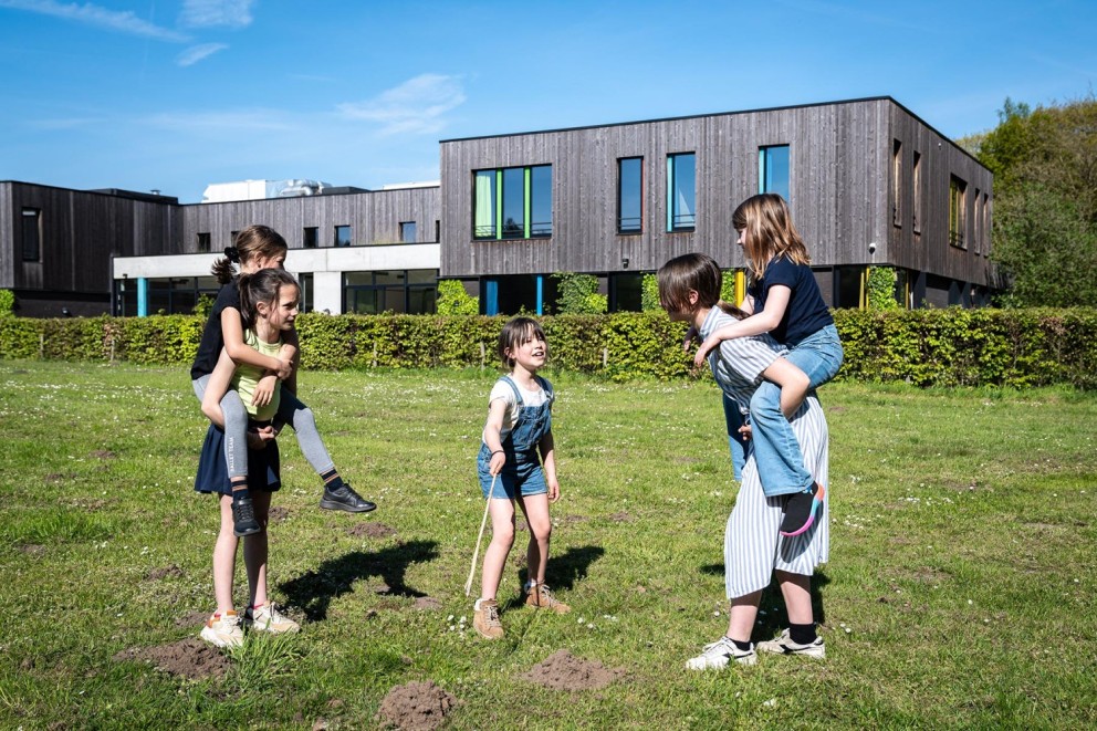 Vijf kinderen spelen buiten in het gras, met twee kinderen op de rug van hun speelgenoten, terwijl de anderen lachen en plezier hebben. Op de achtergrond zijn moderne gebouwen zichtbaar onder een blauwe lucht.