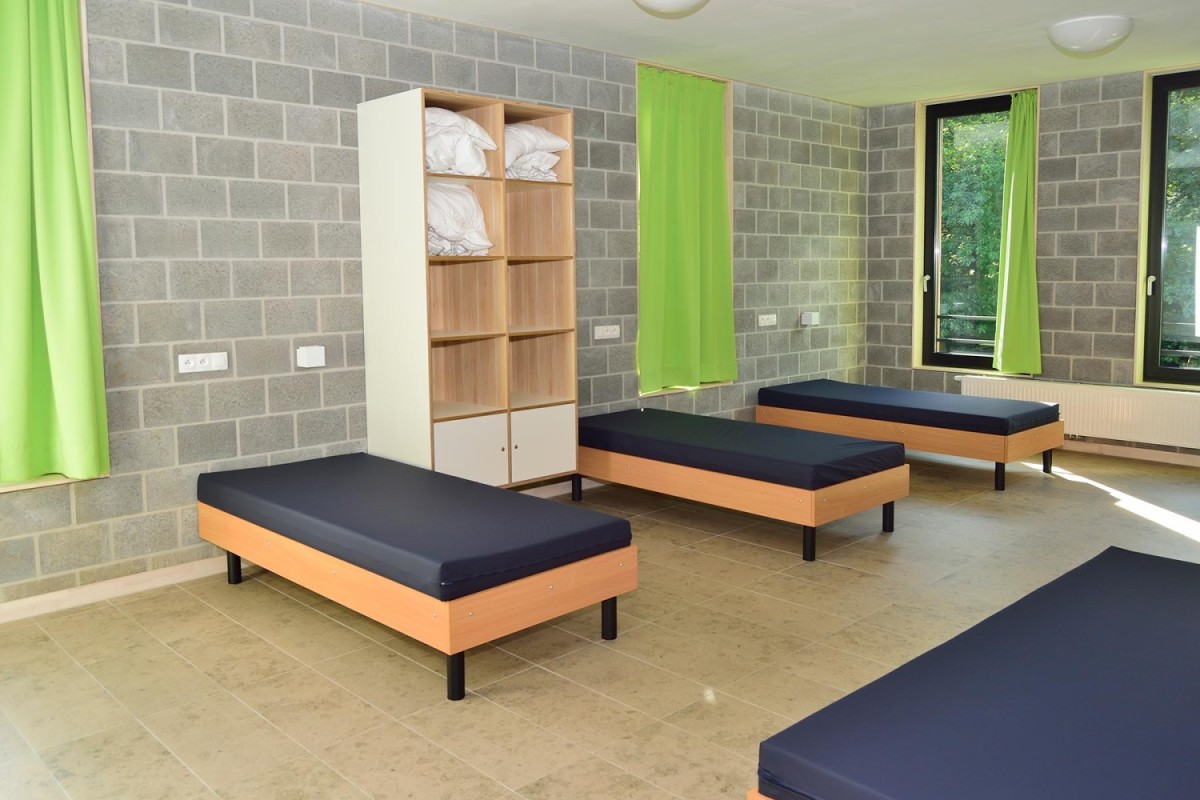 Een eenvoudige kamer met grijze muren, drie bedden met zwarte matrassen, een boekenkast met witte handdoeken en groene gordijnen bij de ramen.