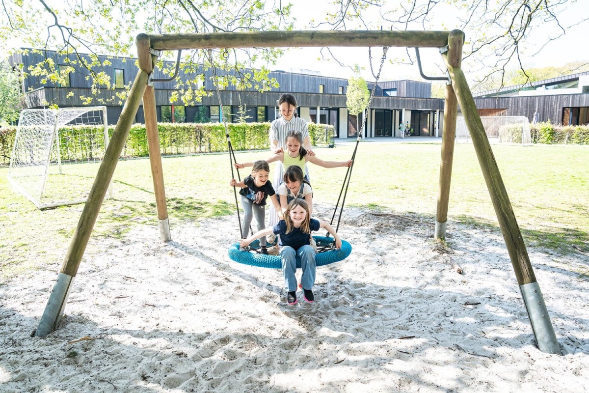Zes kinderen spelen op een schommel in een speeltuin, met een groen grasveld en een gebouw op de achtergrond.