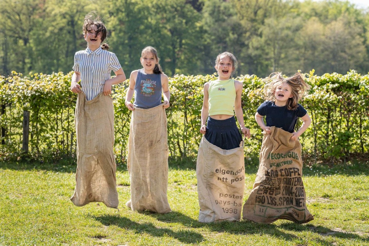 Vier kinderen springen in jute zakken op een groene grasweide, met bomen op de achtergrond en blije gezichten.