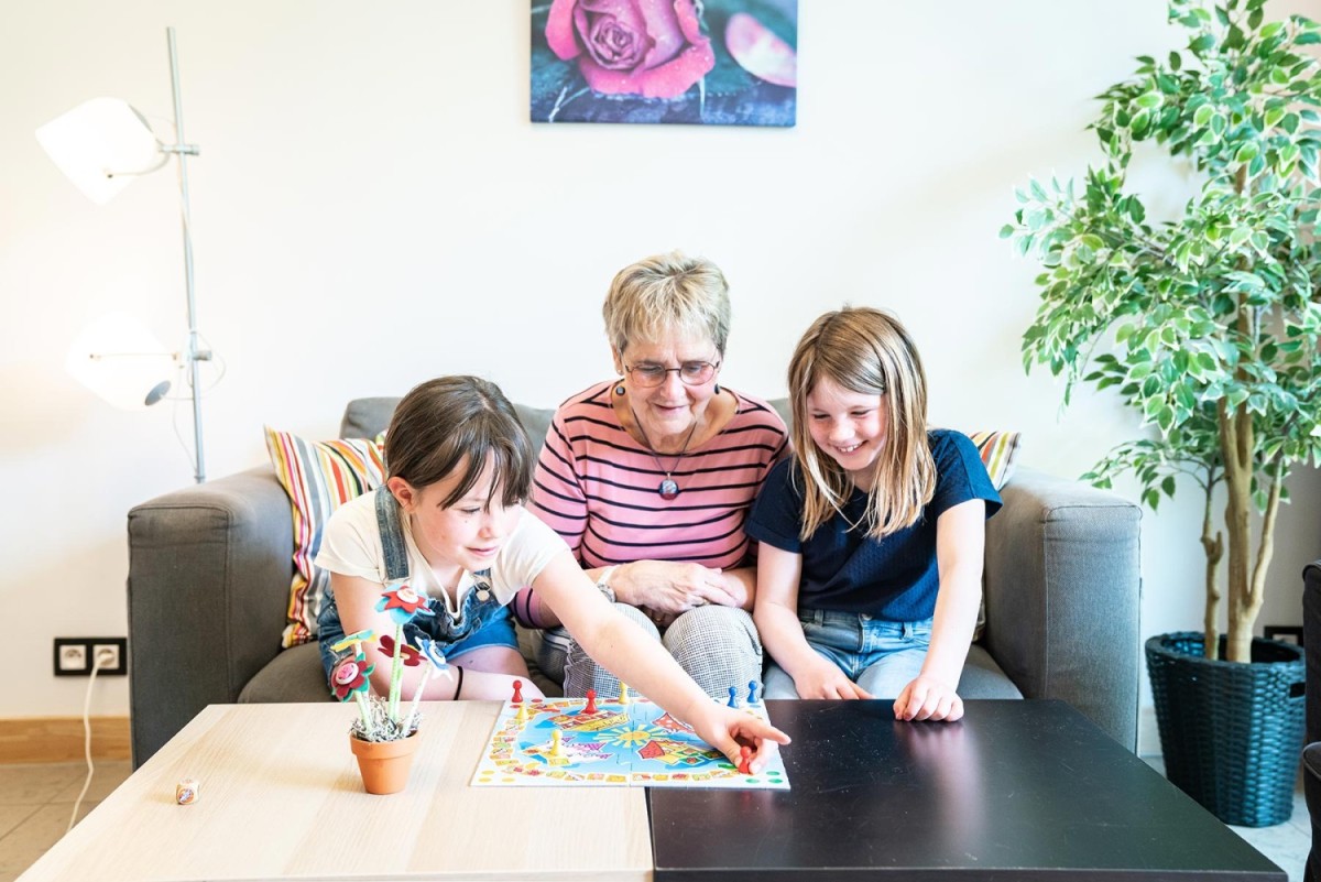Een grootmoeder speelt een bordspel met twee jonge meisjes op een bank, terwijl ze samen lachen en zich concentreren op het spel.