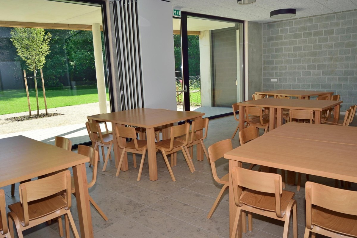 Een moderne eet- of vergaderruimte met houten tafels en stoelen, zicht op een groene tuin door grote ramen.