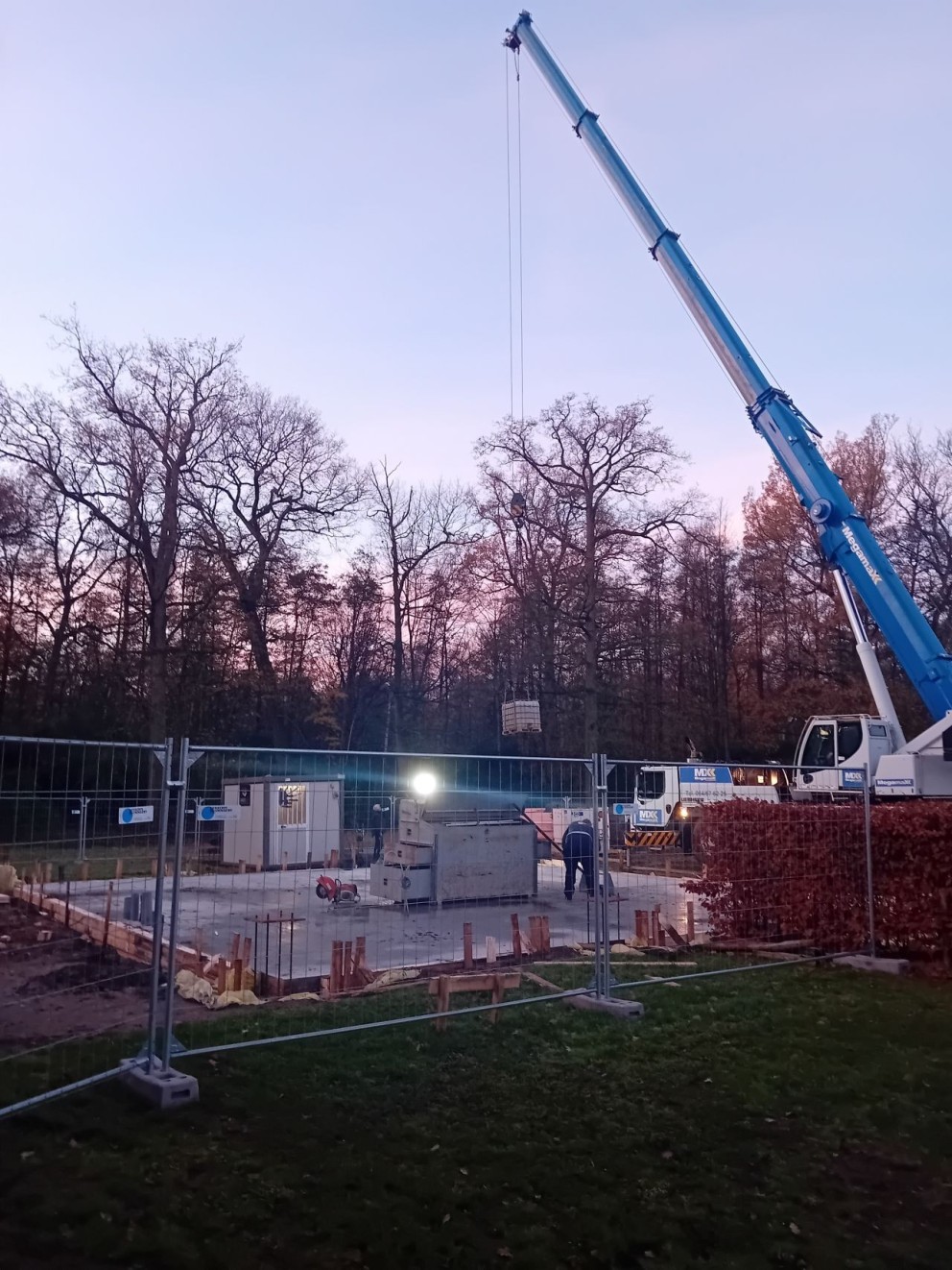 Bouwplaats met een kraan die een object hoog boven een betonfundering heft, omringd door een hek en bomen op de achtergrond tijdens de schemering.