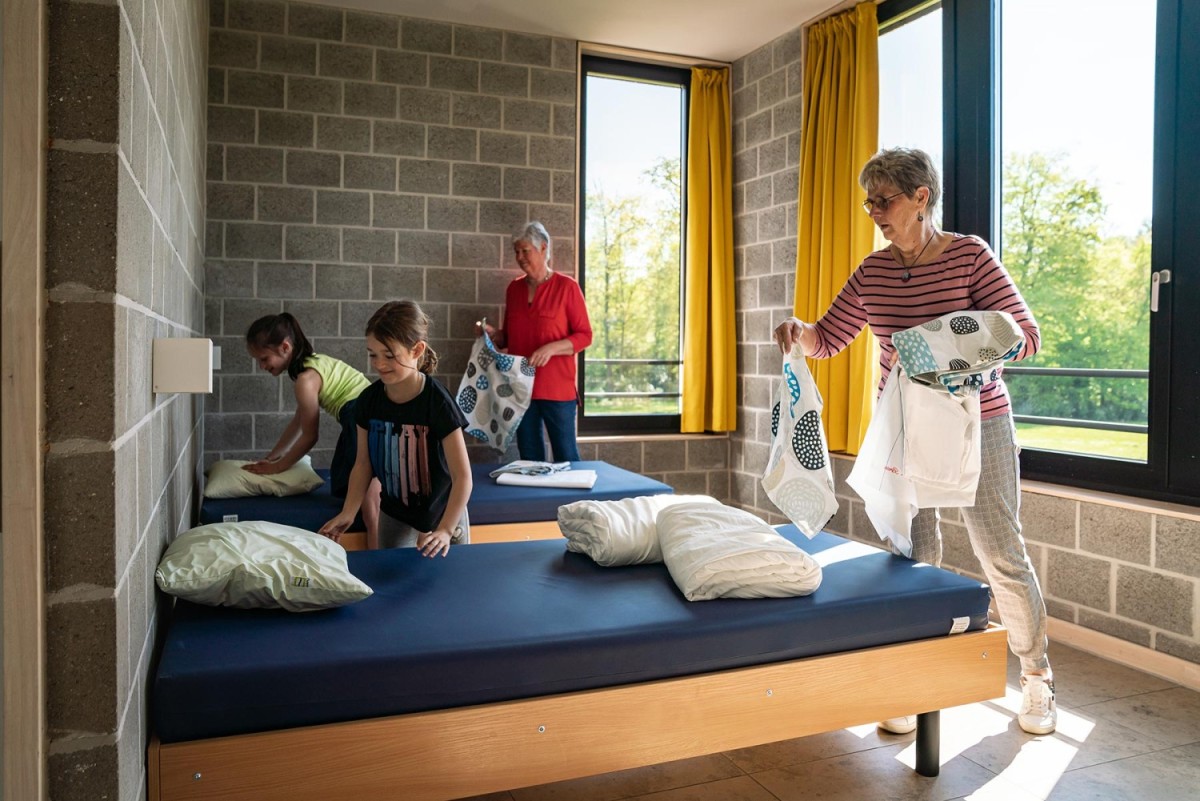 Een groep mensen, waaronder twee kinderen en twee volwassenen, zijn bezig met het opmaken van bedden in een kamer met grote ramen en gele gordijnen.