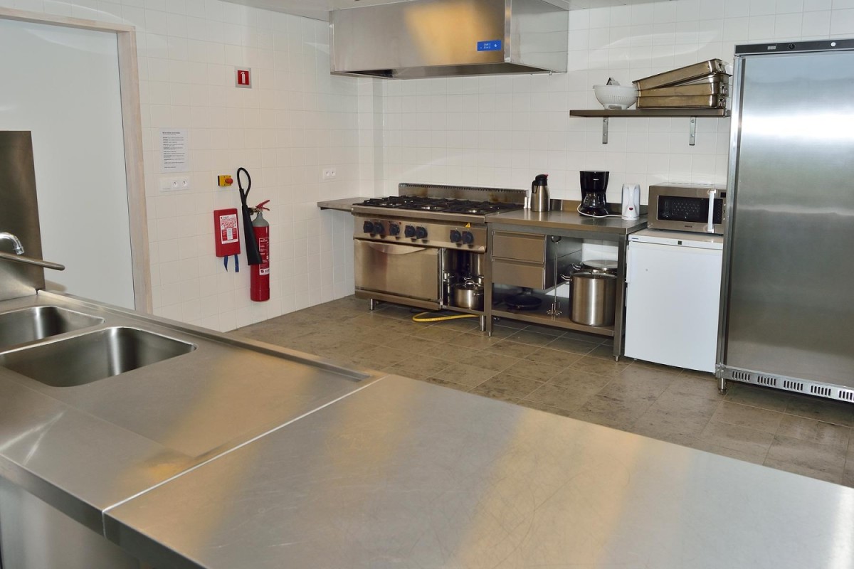 Een moderne, goed uitgeruste keuken met roestvrijstalen werkbladen, kookplaat, oven, magnetron, koffiezetapparaat en een grote koelkast, met een wasbak aan de voorzijde en brandblusser aan de wand.